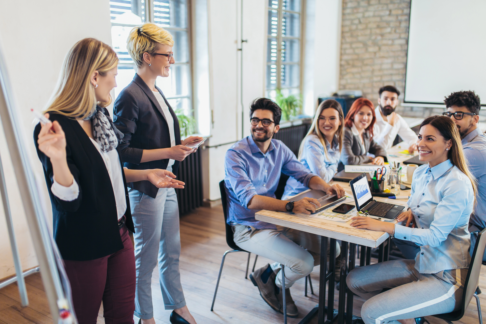 Twee vrouwelijke trainers staan bij een whiteboard terwijl ze presenteren aan een diverse groep van zes professionals die aan een tafel zitten in een moderne vergaderruimte met grote ramen en bakstenen muur. De deelnemers kijken aandachtig en glimlachend naar de presentatie, sommigen hebben laptops voor zich.