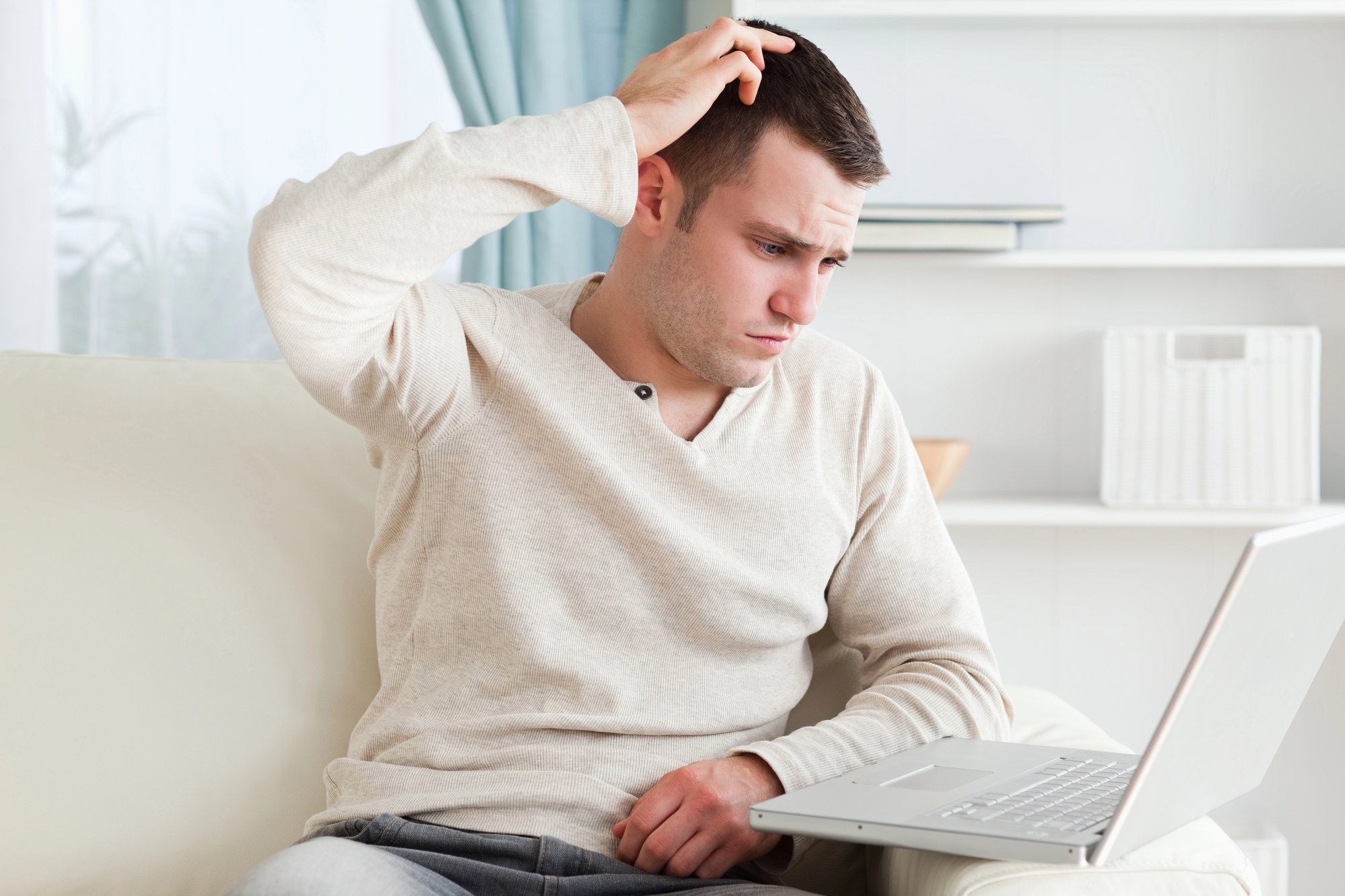 Een jongeman in een beige trui zit op een lichte bank met een laptop op schoot. Hij krabt aan zijn hoofd en kijkt gefrustreerd of verward naar het laptopscherm. De setting is een lichte, moderne kamer met turquoise gordijnen op de achtergrond.